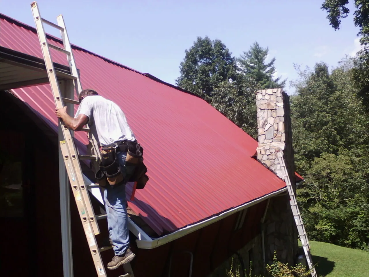 Roofer on a ladder beside a red metal roof and stone chimney — Soffit & Fascia Repair by Roofing Brothers in Radcliff